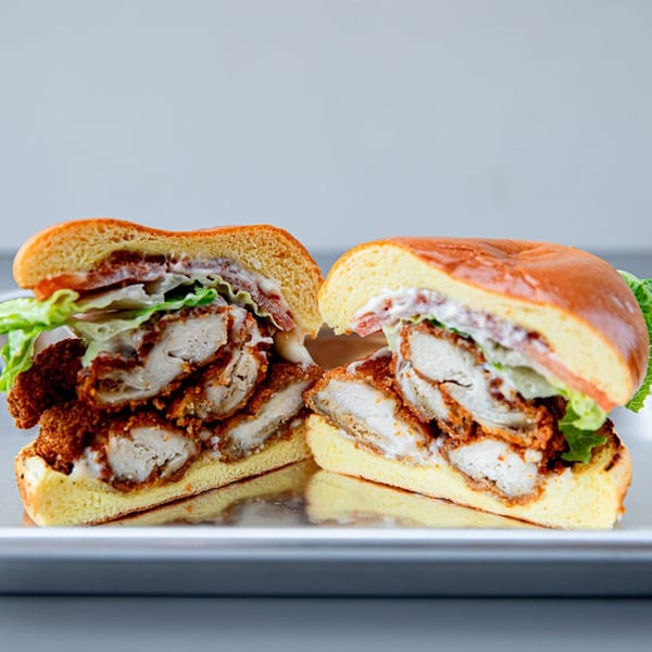 A fried chicken sandwich, cut in half, with lettuce, tomato, and sauce on a brioche bun.